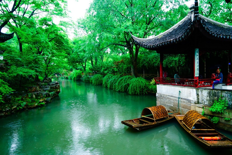 这是一幅中国园林风景图片，图中有青翠的树木，一座亭子，碧绿的池水和两只木船。环境宁静，色彩鲜明。