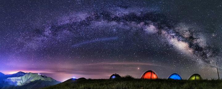 图片展示夜晚露营，几顶彩色帐篷在群山之间，上方是璀璨星河，银河清晰可见，自然美景令人赞叹。