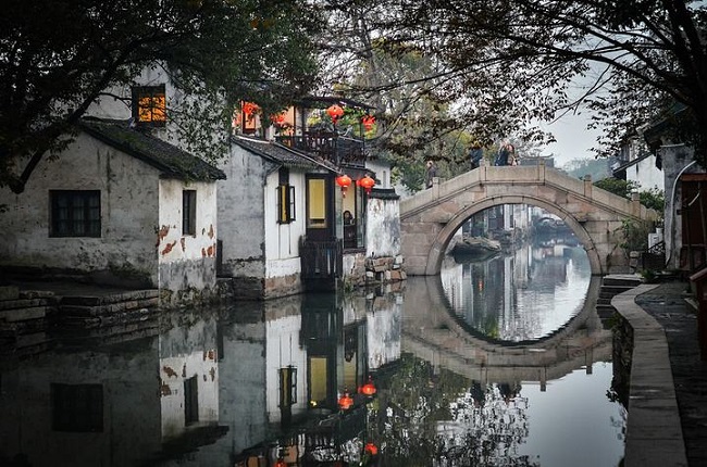 这是一幅中国古镇的照片，照片中有古桥、老式建筑和水面倒影，显出宁静和谐的氛围。红灯笼增添了节日气氛。