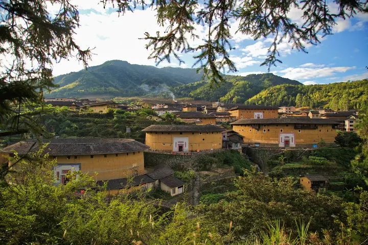 图片展示了一片山区中的土楼群落，楼房圆形，黄土墙面，屋顶瓦片，四周环绕着绿色的树木和山丘。