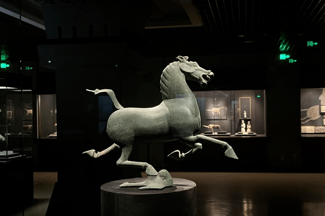 图片展示了一座马的雕塑，四蹄离地，显得生动活泼。背景为昏暗的展厅，几件展品模糊可见。