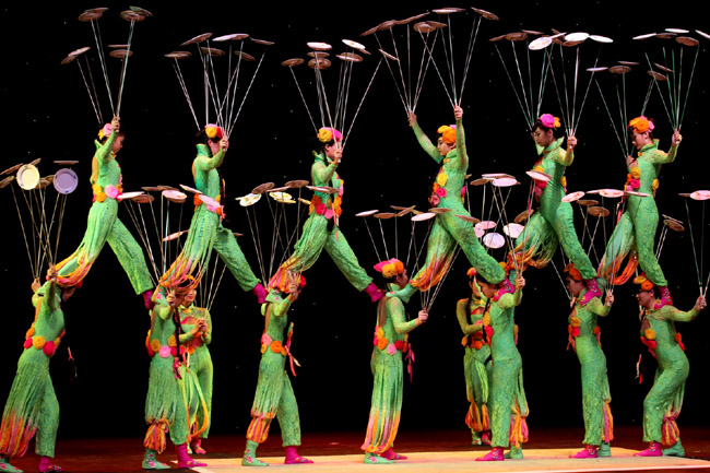 图片展示了身穿绿色服装的表演者在舞台上进行杂技表演，他们头顶和手上旋转着多个盘子。