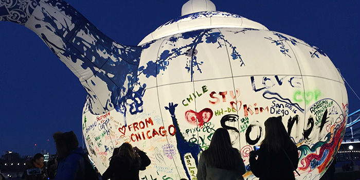 这张图片展示了几个人站在一个巨大的涂鸦球体前，球体上有各种颜色和签名，背景是黄昏或傍晚的天空。