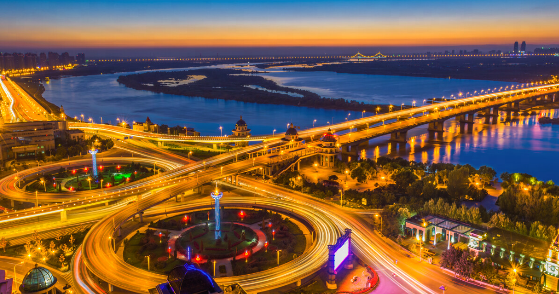 这张图片展示了傍晚时分的城市立交桥，车流灯光形成流动的光带，远处是宁静的河流和天际线，景色宁静而壮观。
