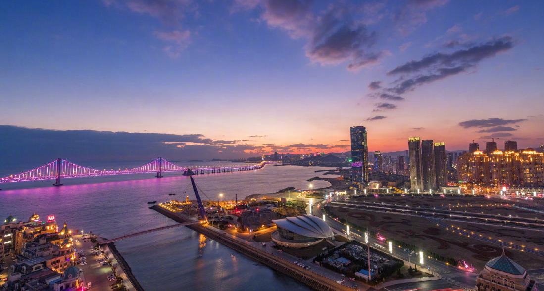 这是一张城市黄昏的风景照片，展示了灯光璀璨的大桥、高楼大厦和宁静的海港，天空中晚霞映照，美丽宁和。