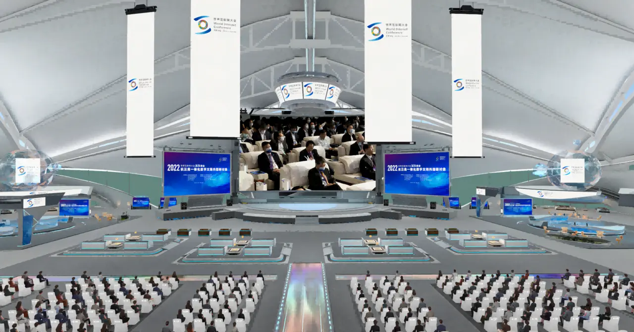 图片展示了一个现代会议室，人们坐着聆听，舞台中央有演讲者，环境科技感强，屏幕显示信息。