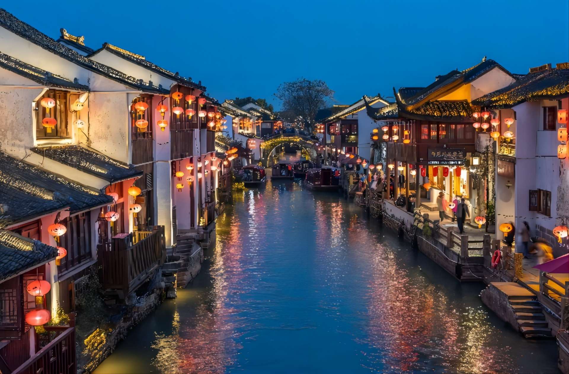 这是一张中国古镇夜景的照片，古建筑沿河而建，红灯笼点缀，水面倒映灯光，游船缓行，营造出宁静而古朴的氛围。