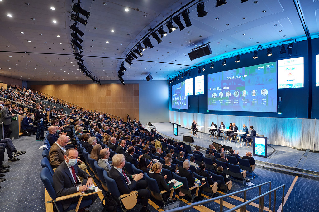 图片展示了一个室内会议场景，台上有发言者，台下观众席坐满了人，大屏幕上显示着演讲内容。