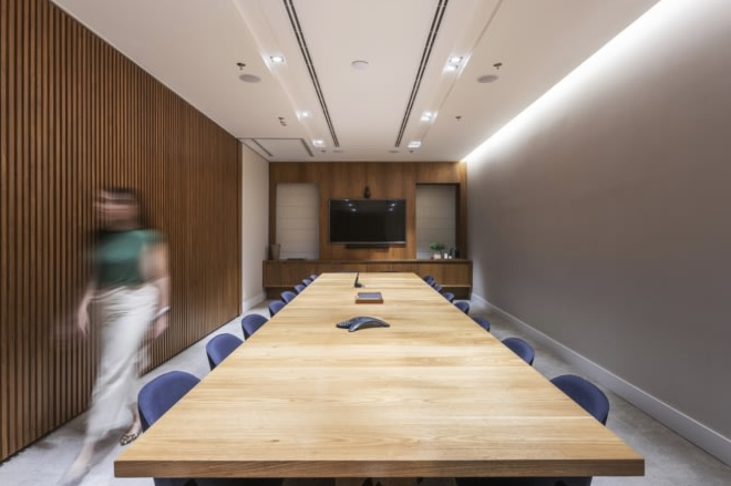 这是一间现代风格的会议室，配有长木质会议桌和蓝色座椅，墙面装饰木质条板，一侧墙上安装有电视。一人模糊行走中。