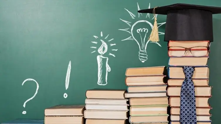 图片展示了一堆书旁边的黑板上画着灯泡和蜡烛，象征灵感和学习，书顶放着学位帽和领带，暗示教育成就。