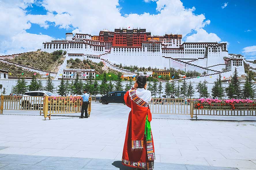 图片展示一位身着藏族传统服饰的人在拍摄布达拉宫，背景是晴朗的天空和远处的雄伟建筑。