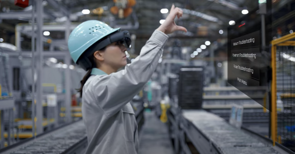 图片中是一位戴着安全头盔和眼镜的女士，在工厂内使用增强现实（AR）技术进行操作或检查工作。