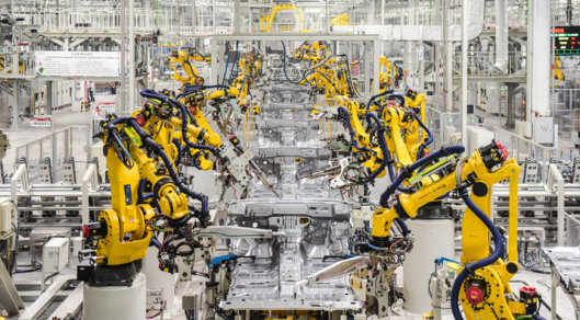 这是一家现代化的汽车制造厂，多个黄色机械臂正在忙碌地进行车辆的组装工作，工厂内部设备先进，布局井然有序。