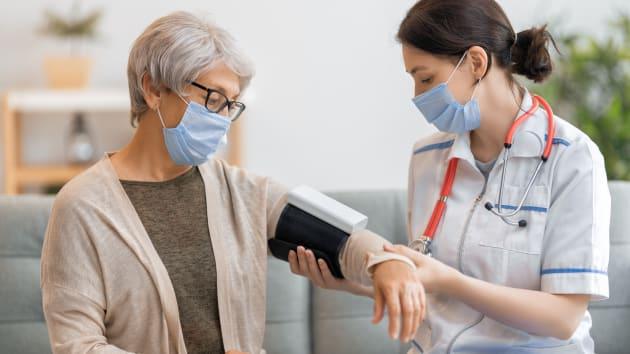 图片展示一位戴口罩的医生在给一位同样戴着口罩的老年女性量血压，两人均穿着便服，背景像家庭环境。