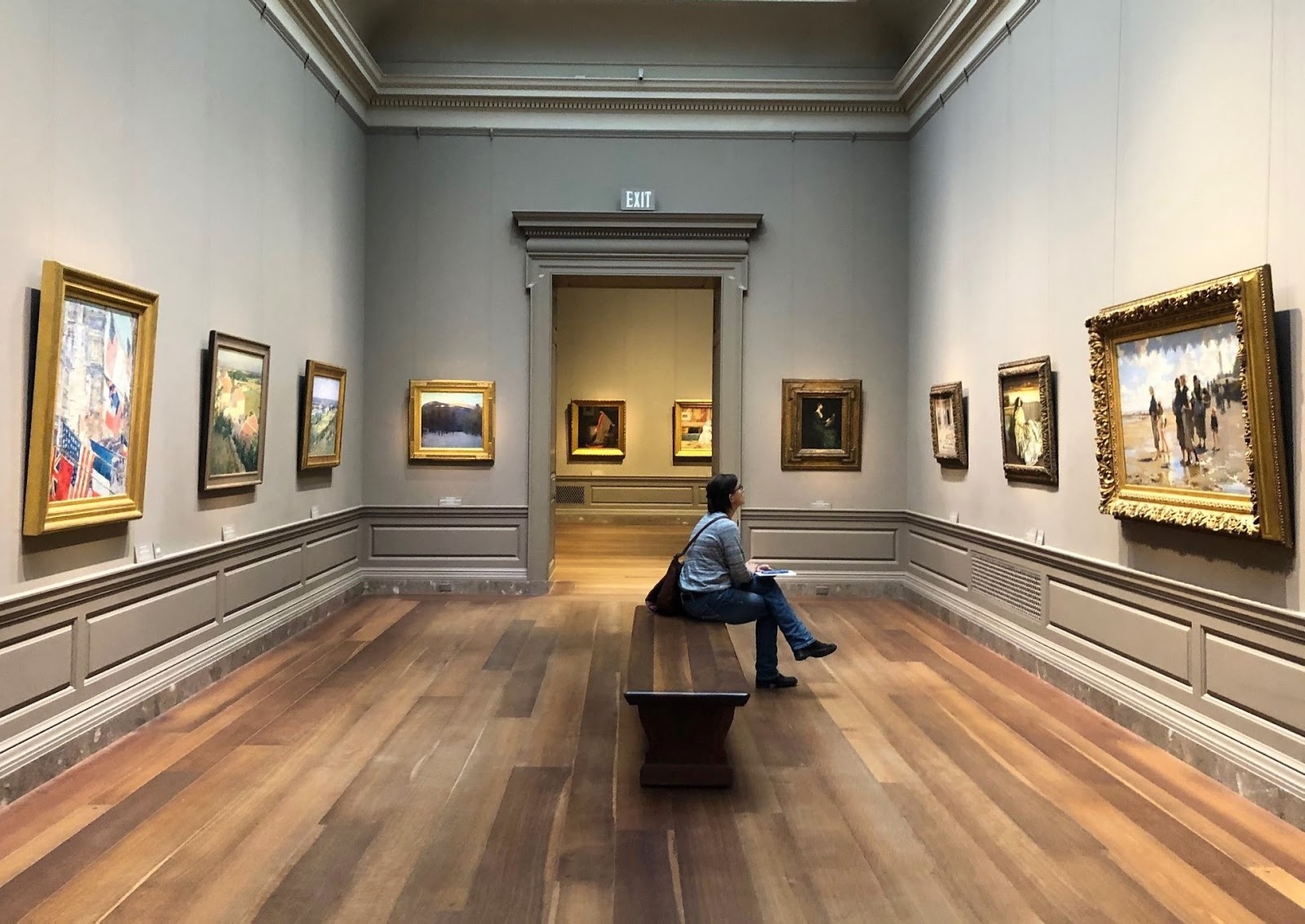 图片展示了一位坐在长椅上的人，身处于装饰典雅的画廊内，墙上挂着多幅精美的画作。