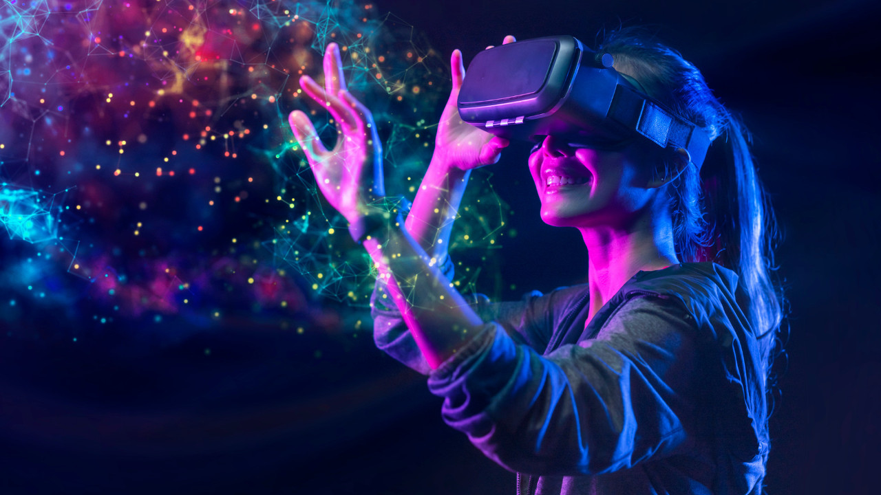 图片展示一位女性佩戴虚拟现实头盔，正兴奋地在光彩斑斓的虚拟空间中伸手触摸，面露笑容。