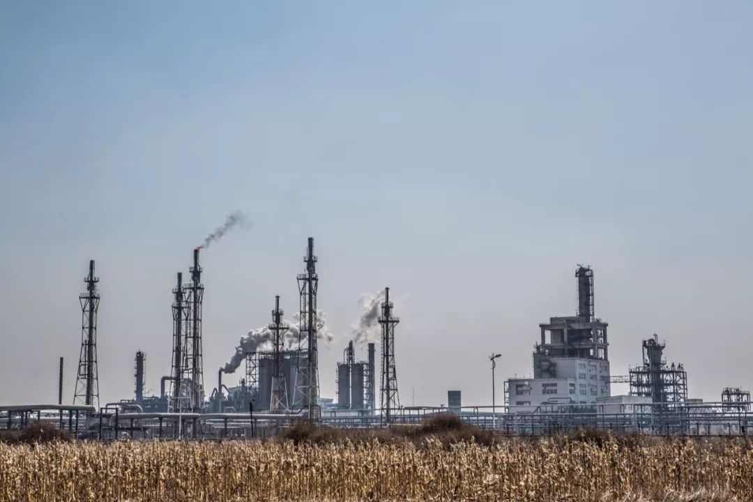 图片显示一座炼油厂，高耸的烟囱排放着烟雾，前景是一片枯黄的草地，体现工业与自然的对比。