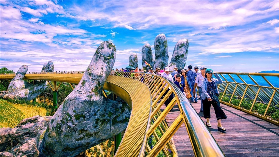 图片展示一座特色桥梁，桥身似巨大手臂支撑，游客在上行走，背景是蓝天白云和宽阔的海景。