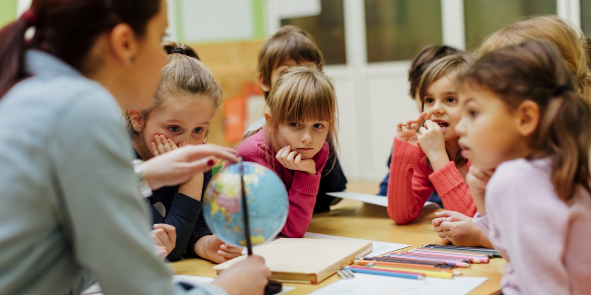 图片展示了一位老师和几个小朋友围坐在桌旁，老师正拿着地球仪向孩子们解释，孩子们表现出专注和好奇的神情。