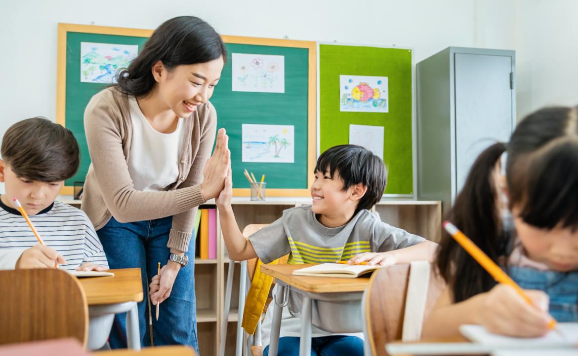 图片展示了一位微笑的女教师在教室里与一个小男孩击掌，周围是专心写作业的其他学生。环境温馨，充满正能量。