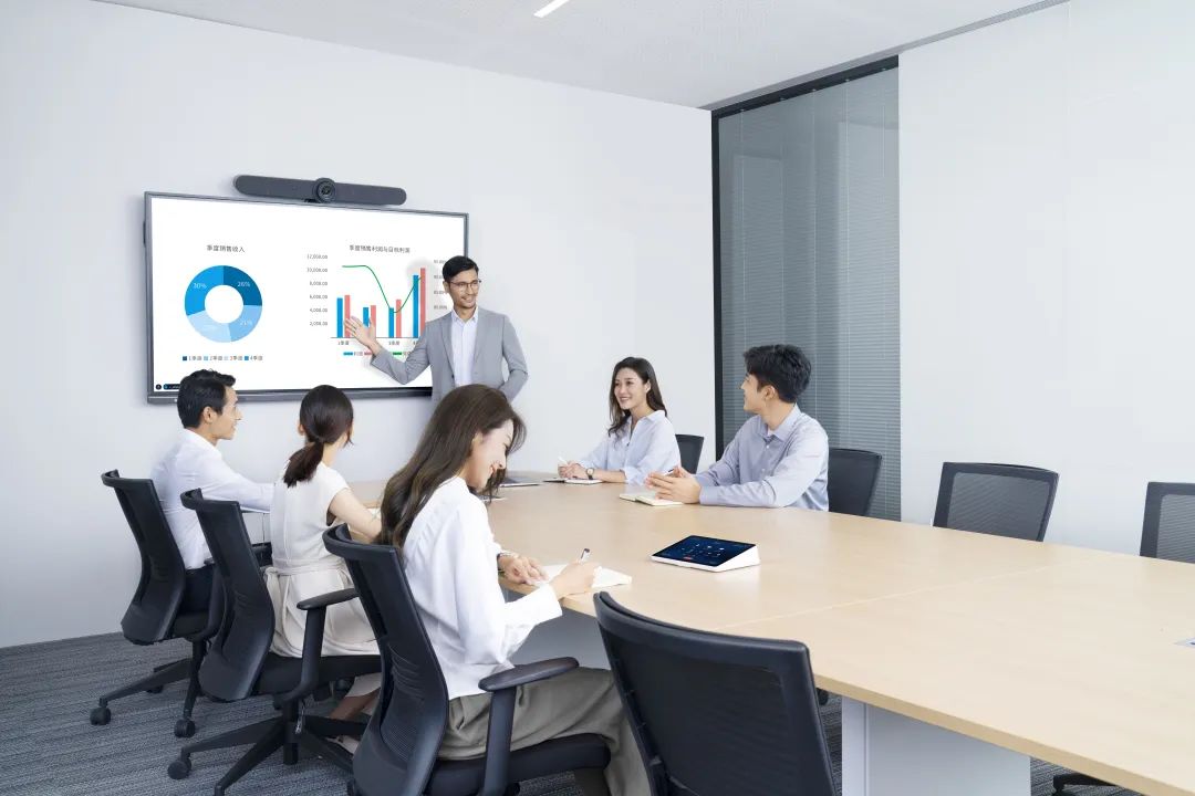 图片展示了一间会议室，里面有一位站立的男士和几位坐着的听众，正在讨论显示在屏幕上的图表和数据。