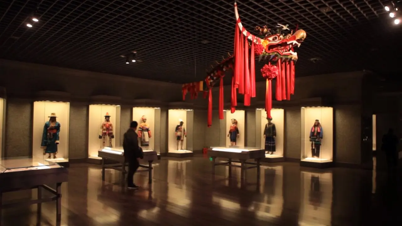 这是一间展览室，墙上挂着红色的中国龙，展示台上陈列着几个穿着传统服饰的人偶，有人正在观看。