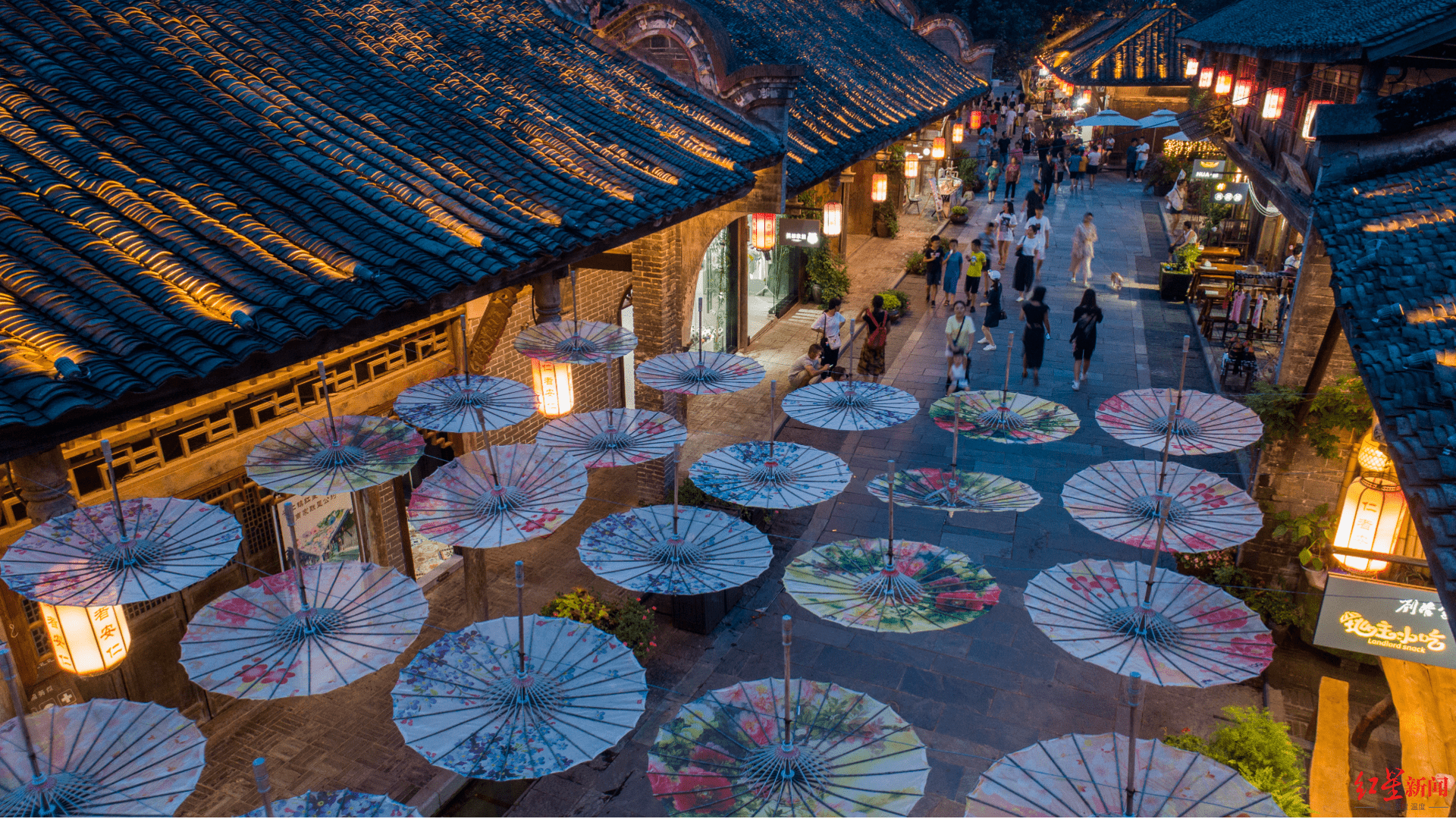 图片展示了一个繁忙的夜市，古老建筑两旁，地面上放置着多把展开的彩色油纸伞，灯光映照下别具风情。