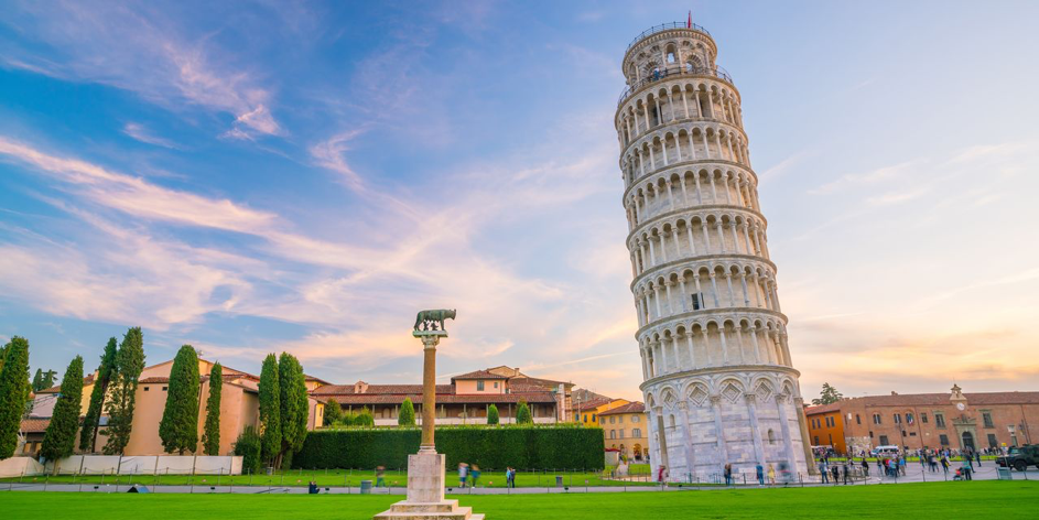 图片展示了意大利著名的比萨斜塔，天空晴朗，塔旁绿草如茵，远处可见建筑群，游客散步在周围。