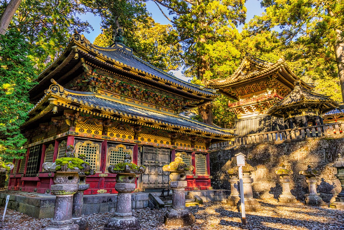 图片展示了一座色彩丰富、装饰精美的日式神社，四周环绕着参天的树木，阳光透过树叶洒在建筑上，显得庄严而宁静。