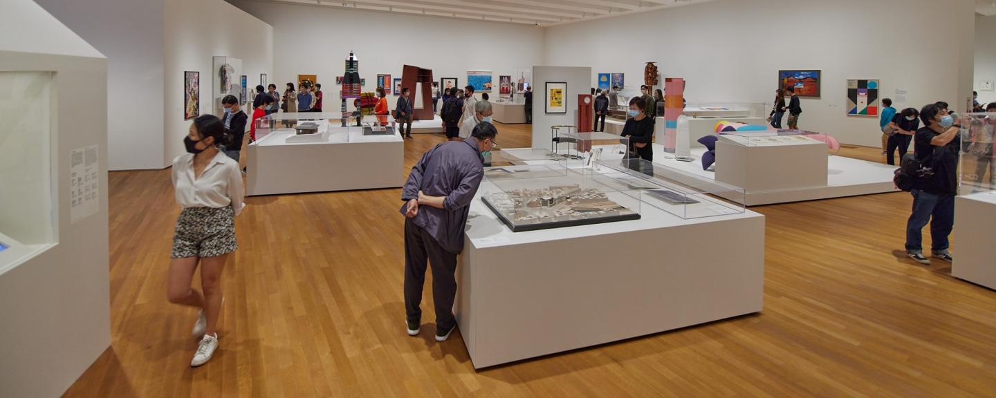 图片展示了一间美术馆内部，多位观众正在欣赏各种展出的艺术品，包括画作和立体作品。环境宽敞明亮。