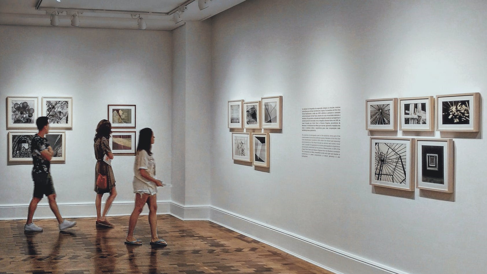 图片展示了三个人在画廊内参观挂在白墙上的不同风格的黑白照片，环境宁静且光线柔和。
