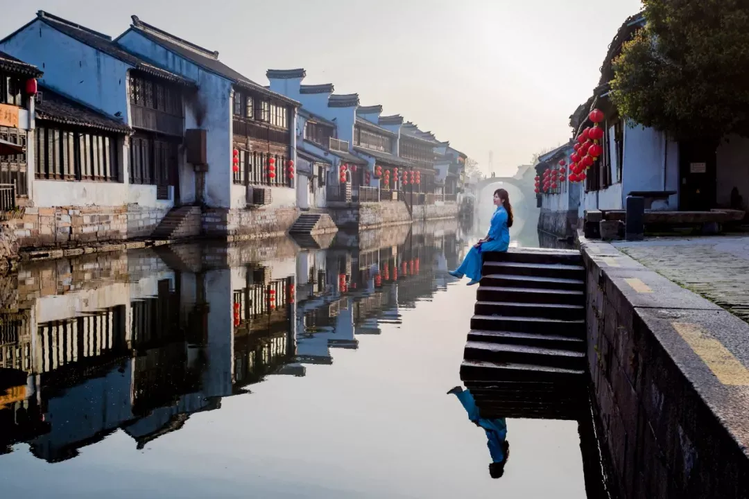 图片展示了一位女士站在古镇的河边，身后是具有中国传统建筑风格的房屋和红灯笼，河面平静反射着周围的景象。