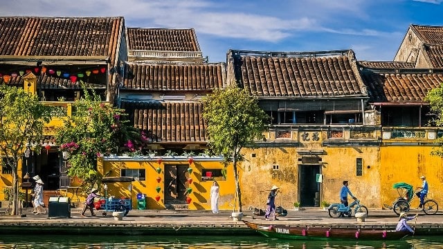 图片展示了亚洲风格的河岸建筑，色彩鲜明，有人骑自行车、划船，体现了宁静与日常生活的融合。