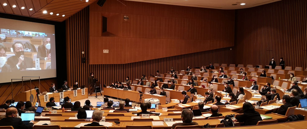 图片展示了一个室内会议场景，多位与会者坐在排列整齐的座椅上，前方是演讲台和大屏幕，屏幕上显示着视频会议的参与者。