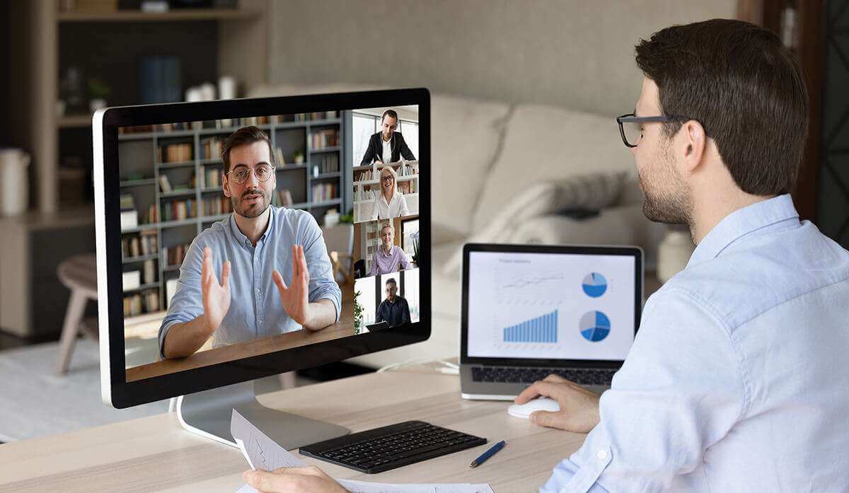 图片展示一位男士正在进行视频会议，电脑屏幕上显示多个参会者，旁边还有一台笔记本电脑显示图表。