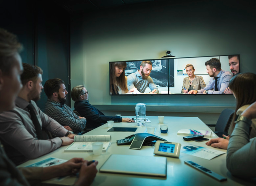 图片展示了一间会议室，几位参会者正坐着，注意力集中在屏幕上的视频会议上，表情专注。会议室内部现代化。