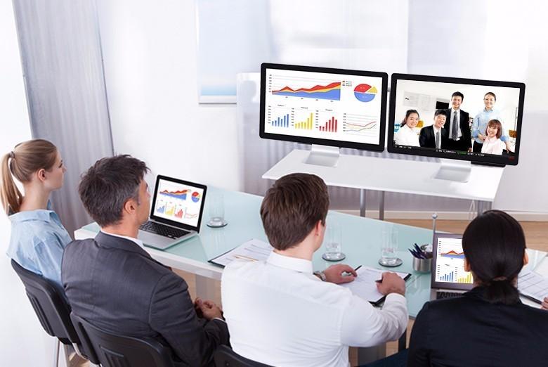 图片展示了四位职场人士正坐在办公室里，通过多显示器视频会议系统与远程同事交流工作事宜。