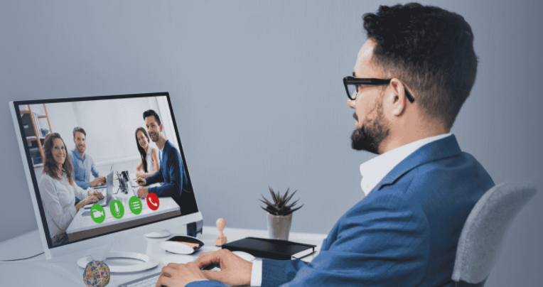 图片展示一位戴眼镜的男士正坐在电脑前，屏幕上显示着几位正在视频会议中的同事。他穿着正装，看起来专注且专业。