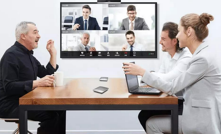 图片展示三人在办公室内进行视频会议，两男一女围坐桌旁，屏幕上显示五位参会者的画面。环境现代，气氛专业。