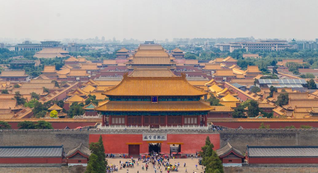 图片展示了故宫的鸟瞰景观，屋顶呈金黄色，周围建筑错落有致，前方聚集了众多游客。天空略显灰蒙。