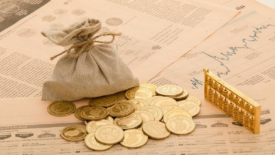 图片展示了一些金币散落在报纸上，旁边有一个布袋和一个金色的金条模型，象征着财富和投资。