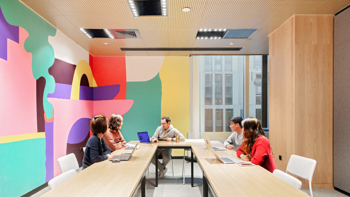 图片展示了四人在现代办公室会议室内开会，墙壁上有彩色抽象图案，环境看起来明亮且色彩丰富。