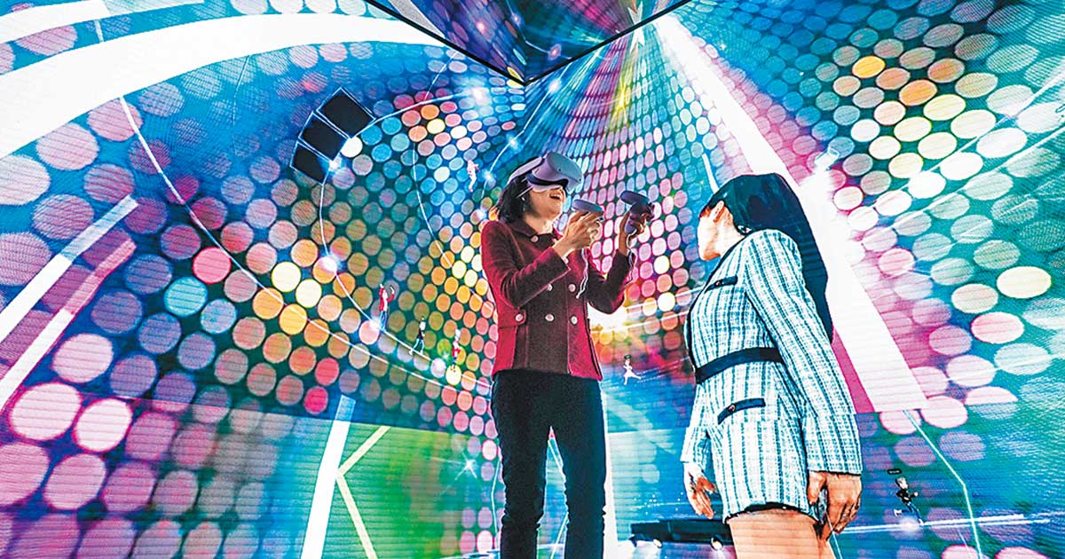 图片展示两人站在充满彩色光点的虚拟现实环境中，一人戴VR头盔，另一人在旁边观看。