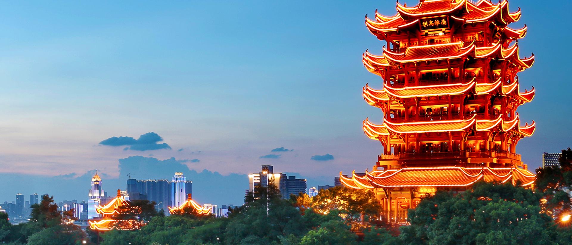 这是一张展示古典中国塔楼在黄昏时分的照片，塔楼灯光亮起，背景是现代城市天际线和树木。