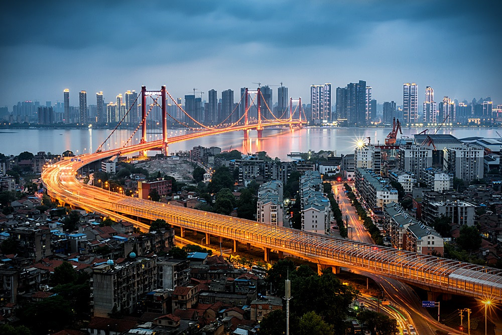 这张图片展示了傍晚时分的城市景观，一座橙色灯光装饰的大桥跨越河流，连接两岸繁华的高楼大厦，车流灯光绘成流动的光带。