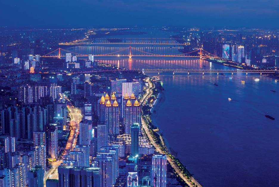 这是一张城市夜景照片，显示了灯光璀璨的高楼大厦，一座大桥跨越宽阔的河流，河面上有船只行驶，远处城市灯火辉煌。
