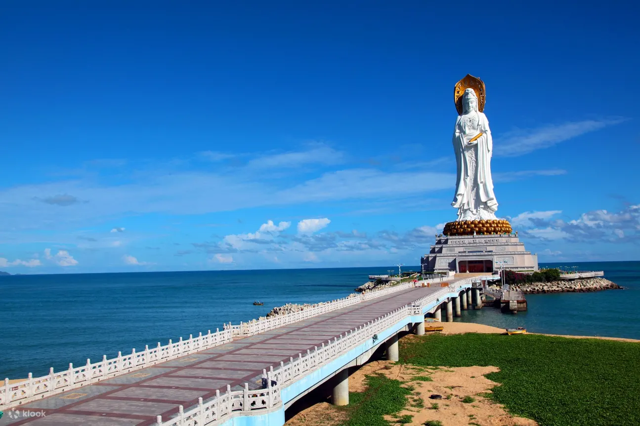 图片展示了一座宏伟的海边观音雕像，前方有一条直通雕像的长桥，背景是蓝天白云和宁静的海洋。