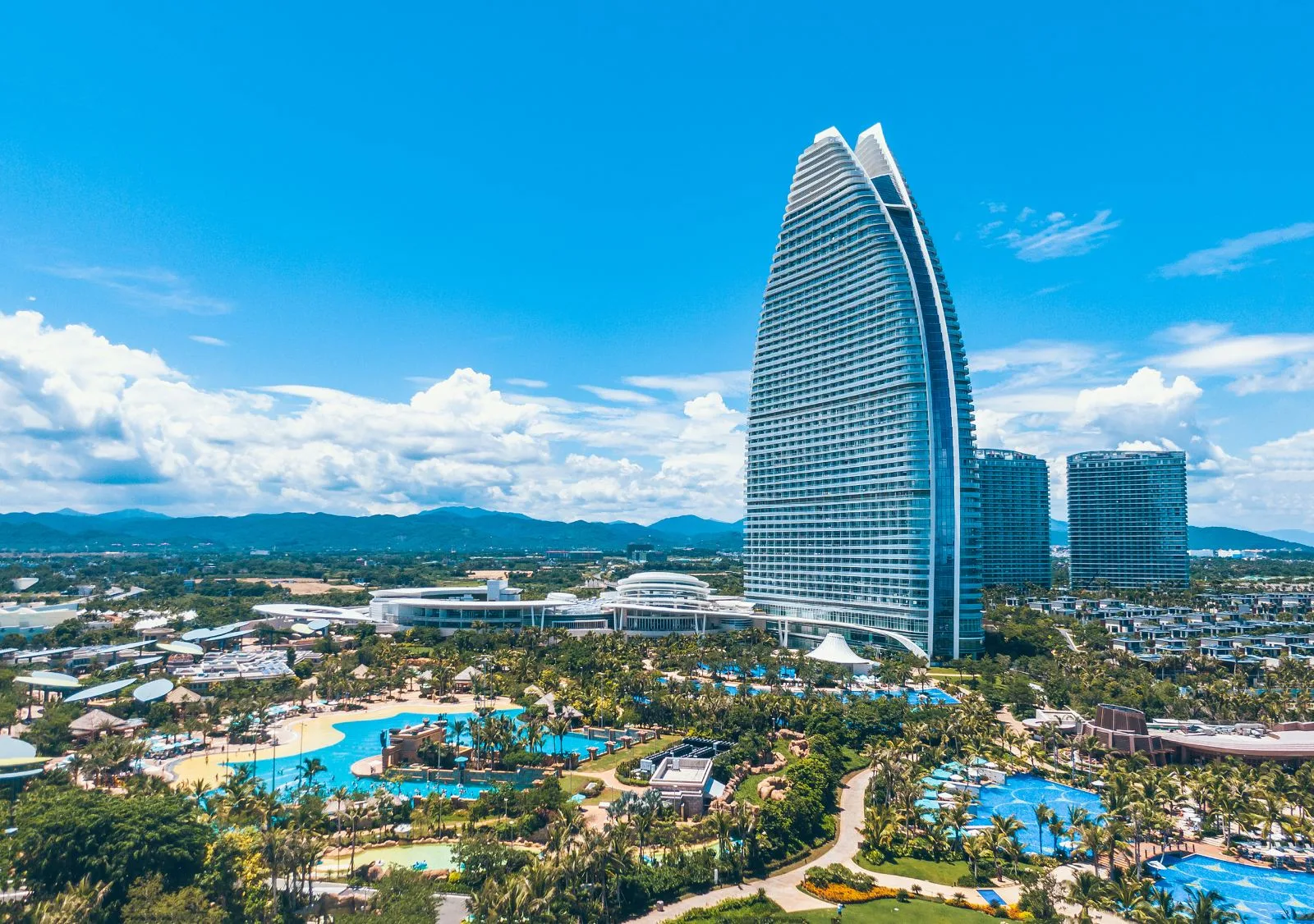 图片展示了一座现代化的高楼大厦，外形似帆船，旁边是热带风光的度假村和碧蓝的游泳池。