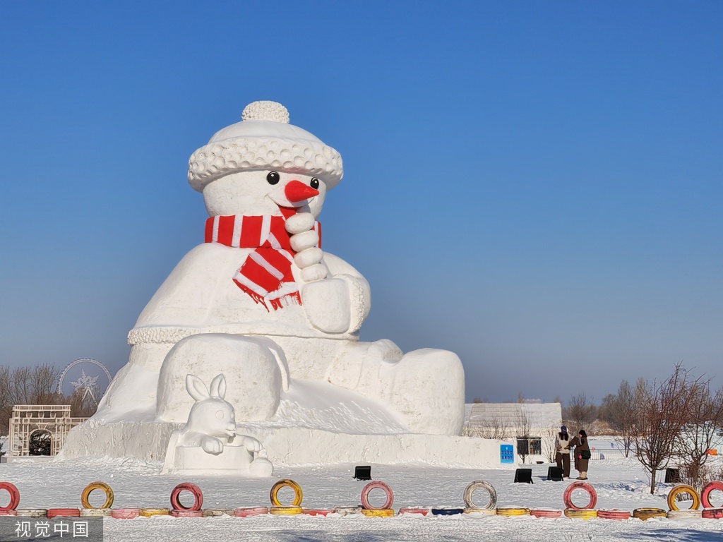 这是一张雪雕图片，展示了一个戴帽子和围巾的大雪人，旁边有一只小雪兔，背景是晴朗的蓝天和几个人。