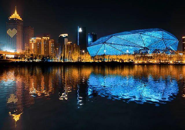 图片展示了夜晚灯光璀璨的现代城市景观，一座独特结构的建筑物在水面上倒映，形成迷人的视觉效果。